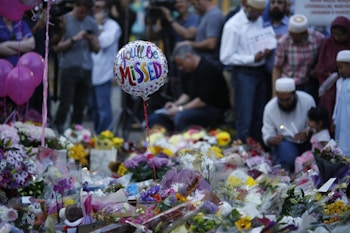 A memorial setup featuring numerous floral tributes and messages. In the foreground, a balloon with the words 'You'll Be Missed' is visible. Several people gather around the site, some holding candles and dressed in diverse attire, suggesting a somber gathering.