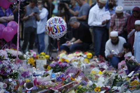 A memorial setup featuring numerous floral tributes and messages. In the foreground, a balloon with the words 'You'll Be Missed' is visible. Several people gather around the site, some holding candles and dressed in diverse attire, suggesting a somber gathering.