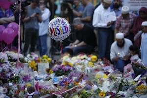 A memorial setup featuring numerous floral tributes and messages. In the foreground, a balloon with the words 'You'll Be Missed' is visible. Several people gather around the site, some holding candles and dressed in diverse attire, suggesting a somber gathering.