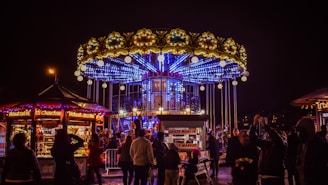 A vibrant carousel with elaborate lighting and decorations is surrounded by a crowd of people at night. The carousel has an upper and lower deck with bright blue neon lights and golden ornamental details. Nearby food stalls and kiosks display various sweets and drinks, illuminated by colorful lights.
