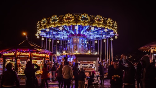 A vibrant carousel with elaborate lighting and decorations is surrounded by a crowd of people at night. The carousel has an upper and lower deck with bright blue neon lights and golden ornamental details. Nearby food stalls and kiosks display various sweets and drinks, illuminated by colorful lights.