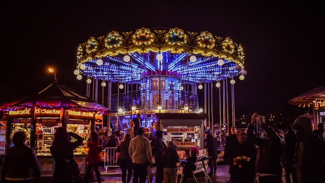 A vibrant carousel with elaborate lighting and decorations is surrounded by a crowd of people at night. The carousel has an upper and lower deck with bright blue neon lights and golden ornamental details. Nearby food stalls and kiosks display various sweets and drinks, illuminated by colorful lights.