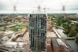 A modern high-rise building is under construction, surrounded by cranes and construction equipment. The scene overlooks a mix of urban and natural landscapes, including houses, roads, trees, and a winding river.