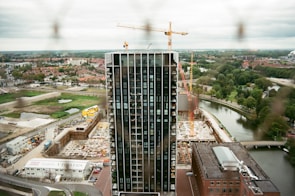 A modern high-rise building is under construction, surrounded by cranes and construction equipment. The scene overlooks a mix of urban and natural landscapes, including houses, roads, trees, and a winding river.