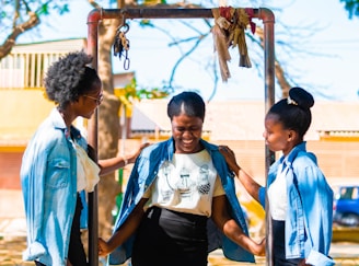 Three women are interacting outdoors in a sunny setting. They are wearing casual outfits with denim jackets. One woman is smiling while being playfully supported by the other two, creating an atmosphere of friendship and joy. A metal structure in the background has some fabric tied to it, and trees and buildings can be seen further back.