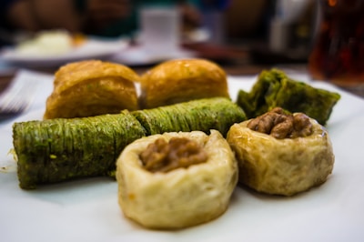 An assortment of baklava pieces arranged on a plate, featuring various types of pastries with flaky textures and different fillings. Some pieces have a visible green hue, indicating pistachio filling, while others have crisp, golden-brown layers. A walnut-topped pastry is also present.