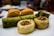 An assortment of baklava pieces arranged on a plate, featuring various types of pastries with flaky textures and different fillings. Some pieces have a visible green hue, indicating pistachio filling, while others have crisp, golden-brown layers. A walnut-topped pastry is also present.