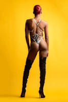 A person with red hair is wearing a strappy one-piece swimsuit and high black heeled boots, standing against a bright yellow background. The pose is from the back, with emphasis on the intricate design of the swimwear.