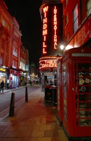 A vibrant street scene at night featuring a red neon sign for 'Windmill International' and 'Table Dancing'. The street is lined with buildings, including a traditional red British telephone booth. Several people are visible walking along the sidewalk.