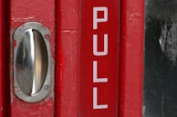 A red door with a metal handle and the word 'PULL' printed in large white letters. The metal handle is shiny and mounted on the left side of the door. The surface of the door shows some signs of wear, with small patches of paint missing near the handle.
