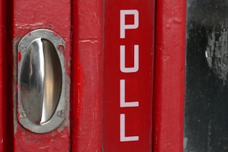 A red door with a metal handle and the word 'PULL' printed in large white letters. The metal handle is shiny and mounted on the left side of the door. The surface of the door shows some signs of wear, with small patches of paint missing near the handle.