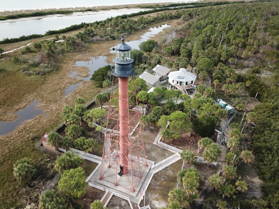 A red lighthouse stands amidst lush green foliage, surrounded by trees and marshland. Adjacent to the lighthouse, there is a building with solar panels on its roof. The landscape extends to a distant shoreline.