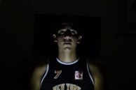 A person is central in the foreground with dim lighting creating a shadowed, dramatic effect. The person is wearing a black sports jersey with the word 'New York' visible and the number 7. Most of the background is dark, enhancing the focus on the illuminated face and upper torso.