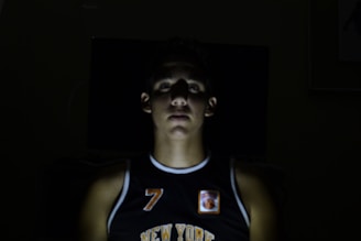A person is central in the foreground with dim lighting creating a shadowed, dramatic effect. The person is wearing a black sports jersey with the word 'New York' visible and the number 7. Most of the background is dark, enhancing the focus on the illuminated face and upper torso.