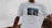 A person wearing a white T-shirt featuring an image of two hands reaching towards the sky, surrounded by palm trees. The background is slightly blurred, focusing on the T-shirt's design.
