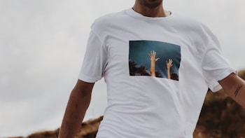 A person wearing a white T-shirt featuring an image of two hands reaching towards the sky, surrounded by palm trees. The background is slightly blurred, focusing on the T-shirt's design.