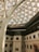 A modern architectural interior features a geometric glass dome ceiling supported by large metal beams. The building has a white facade with multiple rectangular windows, and there is a large artwork on one of the walls depicting a bird.