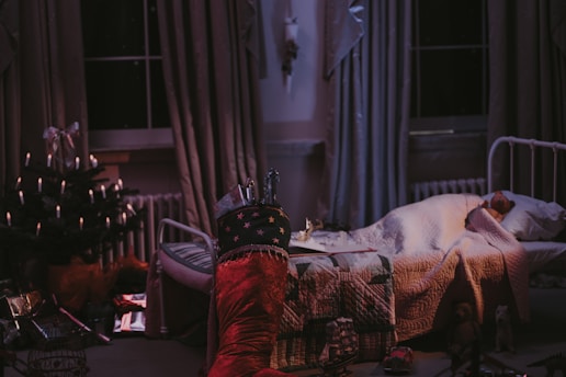 A cozy bedroom with a sleeping child tucked under a quilted blanket. A festive atmosphere is suggested by a small Christmas tree adorned with candles and presents under it. Stockings are hung, one filled with gifts. The room is softly lit, suggesting a peaceful nighttime setting.