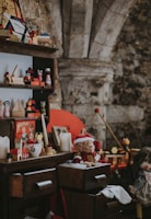 A vintage scene featuring a wooden shelf and dresser filled with assorted nostalgic toys and decorations. Items include a teddy bear wearing a Santa hat, a vintage board game box, candles, and various wooden toys. The setting seems to evoke a cozy, old-fashioned atmosphere with a rustic stone wall in the background.