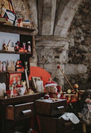 A vintage scene featuring a wooden shelf and dresser filled with assorted nostalgic toys and decorations. Items include a teddy bear wearing a Santa hat, a vintage board game box, candles, and various wooden toys. The setting seems to evoke a cozy, old-fashioned atmosphere with a rustic stone wall in the background.