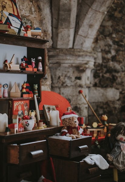 A vintage scene featuring a wooden shelf and dresser filled with assorted nostalgic toys and decorations. Items include a teddy bear wearing a Santa hat, a vintage board game box, candles, and various wooden toys. The setting seems to evoke a cozy, old-fashioned atmosphere with a rustic stone wall in the background.