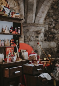 A vintage scene featuring a wooden shelf and dresser filled with assorted nostalgic toys and decorations. Items include a teddy bear wearing a Santa hat, a vintage board game box, candles, and various wooden toys. The setting seems to evoke a cozy, old-fashioned atmosphere with a rustic stone wall in the background.