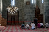 A group of people is engaged in prayer within a mosque characterized by intricate architectural details. The interior features ornate wooden carvings, a red patterned carpet, and a large brass chandelier. The walls and decor include Arabic script, adding to the spiritual ambiance of the setting.