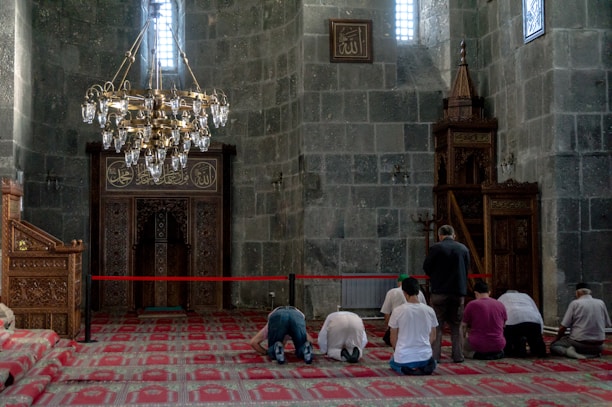 A group of people is engaged in prayer within a mosque characterized by intricate architectural details. The interior features ornate wooden carvings, a red patterned carpet, and a large brass chandelier. The walls and decor include Arabic script, adding to the spiritual ambiance of the setting.