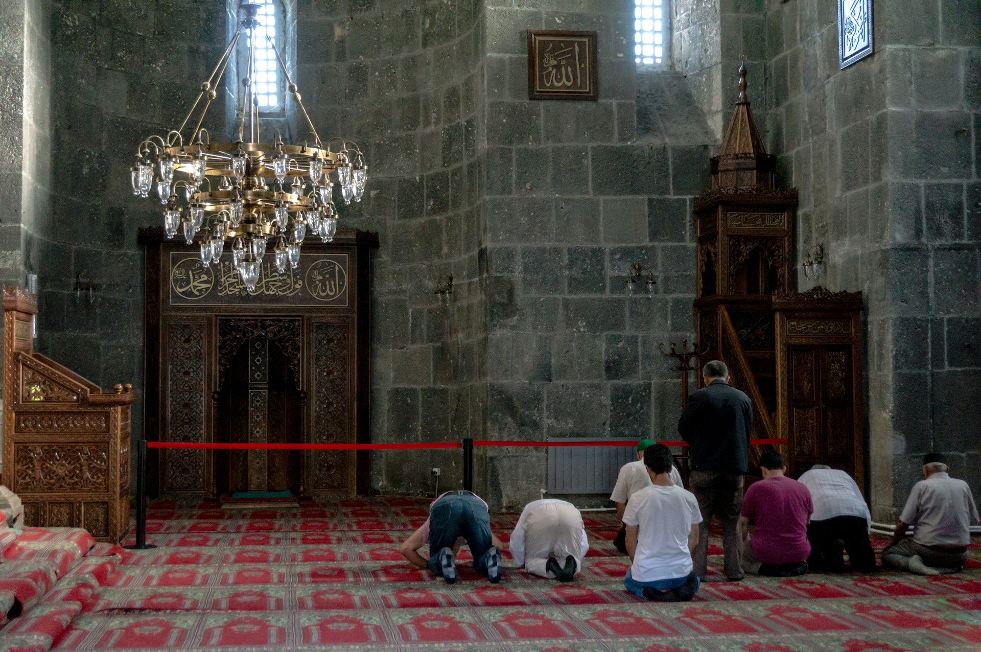 A group of people is engaged in prayer within a mosque characterized by intricate architectural details. The interior features ornate wooden carvings, a red patterned carpet, and a large brass chandelier. The walls and decor include Arabic script, adding to the spiritual ambiance of the setting.