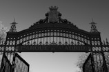 An ornate wrought iron gate with intricate patterns and a crest at the top. The words 'PARQUE de MADRID' are prominently displayed in the center of the gate. The gate is set against a clear sky and is flanked by trees on either side.