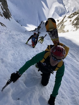 Three climbers wearing winter gear are ascending a steep, snow-covered slope between two rock faces. The lead climber uses an ice axe, and skis are strapped to their backpack. The other two climbers follow closely behind, using similar climbing gear.