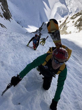 Three climbers wearing winter gear are ascending a steep, snow-covered slope between two rock faces. The lead climber uses an ice axe, and skis are strapped to their backpack. The other two climbers follow closely behind, using similar climbing gear.