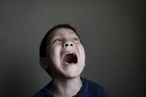 A young boy with short dark hair is opening his mouth wide, possibly screaming or shouting. The lighting is subdued, with a dark background that contrasts with his lighter skin tone. His expression conveys strong emotion, possibly excitement or frustration.