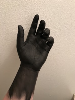 A hand is covered in black paint against a textured, light-colored wall. The fingers are slightly curled, and the paint appears smooth and glossy.