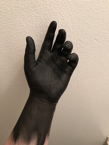A hand is covered in black paint against a textured, light-colored wall. The fingers are slightly curled, and the paint appears smooth and glossy.