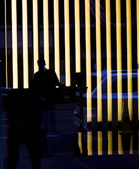 Vertical yellow light bars create a striking pattern. A silhouette of a person is visible in the foreground, adding a mysterious element. Reflections of a vehicle and street elements can be seen, contributing to an urban atmosphere.