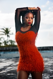 A woman stands confidently with hands raised above her head, wearing a vibrant orange textured dress. Her bold red lipstick and braided hairstyle complement her elegant look. The background features a blurred view of trees and a rooftop under a partly cloudy sky.