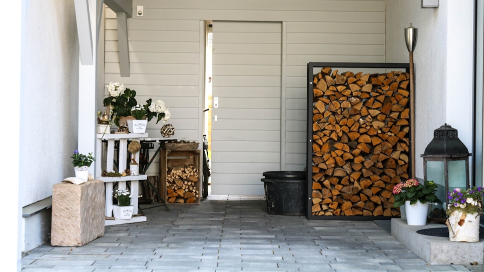 A neatly organized outdoor area with stacked firewood framed in a modern container against a light-colored wooden wall. Various potted plants, including colorful flowers and greenery, are arranged on white shelves and the ground. The space also features a rustic stone block and a decorative lantern, creating a welcoming and cozy atmosphere.