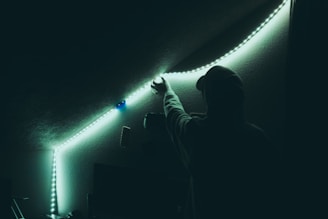 A person wearing a hoodie reaches up to adjust a strip of LED lights along the ceiling of a dimly lit room. The lights cast a soft, greenish-blue glow on the surrounding walls.