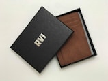 A brown leather wallet is partially visible inside an open black box. The box has a textured surface with the letters 'RVI' embossed in white. The wallet features multiple card slots and appears to be of high quality.