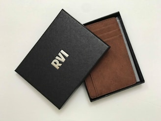 A brown leather wallet is partially visible inside an open black box. The box has a textured surface with the letters 'RVI' embossed in white. The wallet features multiple card slots and appears to be of high quality.