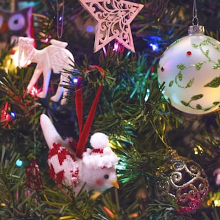 A decorated Christmas tree adorned with various ornaments, including a carved wooden star, a white camel, and a plush bird wearing a knitted outfit. There are also colorful lights and a large round bauble with green vine patterns.