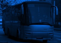 A large bus is parked on the side of the road, partially obscured by surrounding trees. The image is tinted in a monochromatic blue. The bus has a 'Tourist' sign displayed on the front window, and the driver is visible inside.