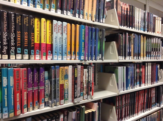 Rows of colorful books neatly arranged on white library shelves. The spines display various titles and authors, reflecting diverse genres. The arrangement suggests organization and accessibility for readers.