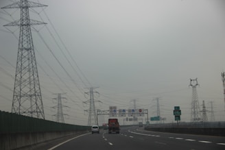 A highway with vehicles traveling on it is flanked by a series of tall power transmission towers. Overhead, the sky is overcast and grey, creating a subdued atmosphere. Road signs with Chinese characters are visible above the road, and there are several lanes suggesting a busy route.