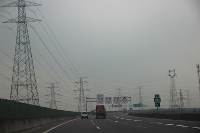 A highway with vehicles traveling on it is flanked by a series of tall power transmission towers. Overhead, the sky is overcast and grey, creating a subdued atmosphere. Road signs with Chinese characters are visible above the road, and there are several lanes suggesting a busy route.