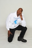 A person is kneeling on one knee, wearing a white hoodie with a blue earth emblem and the words 'Heaven and Earth'. The individual is looking upward with a thoughtful expression, with their right hand touching their face. They are dressed in black pants and black shoes.