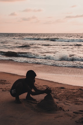 A child crouches on a sandy beach, intently building a sandcastle as gentle waves roll onto the shore under a warm, glowing sky at sunset.