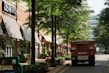 A shopping street with several storefronts, including a DSW Shoes store with black and white striped awnings. Trees line the walkway, providing shade. There is a red armored truck parked on the street and people can be seen walking or sitting on benches along the sidewalk. The surrounding area appears to be clean and well-maintained, with street lights and potted plants along the path.