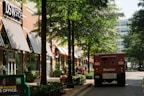 A shopping street with several storefronts, including a DSW Shoes store with black and white striped awnings. Trees line the walkway, providing shade. There is a red armored truck parked on the street and people can be seen walking or sitting on benches along the sidewalk. The surrounding area appears to be clean and well-maintained, with street lights and potted plants along the path.